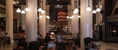 A spacious lobby with high ceilings, large columns, and hanging pendant lights. There is a bar area in the background with a large American flag.