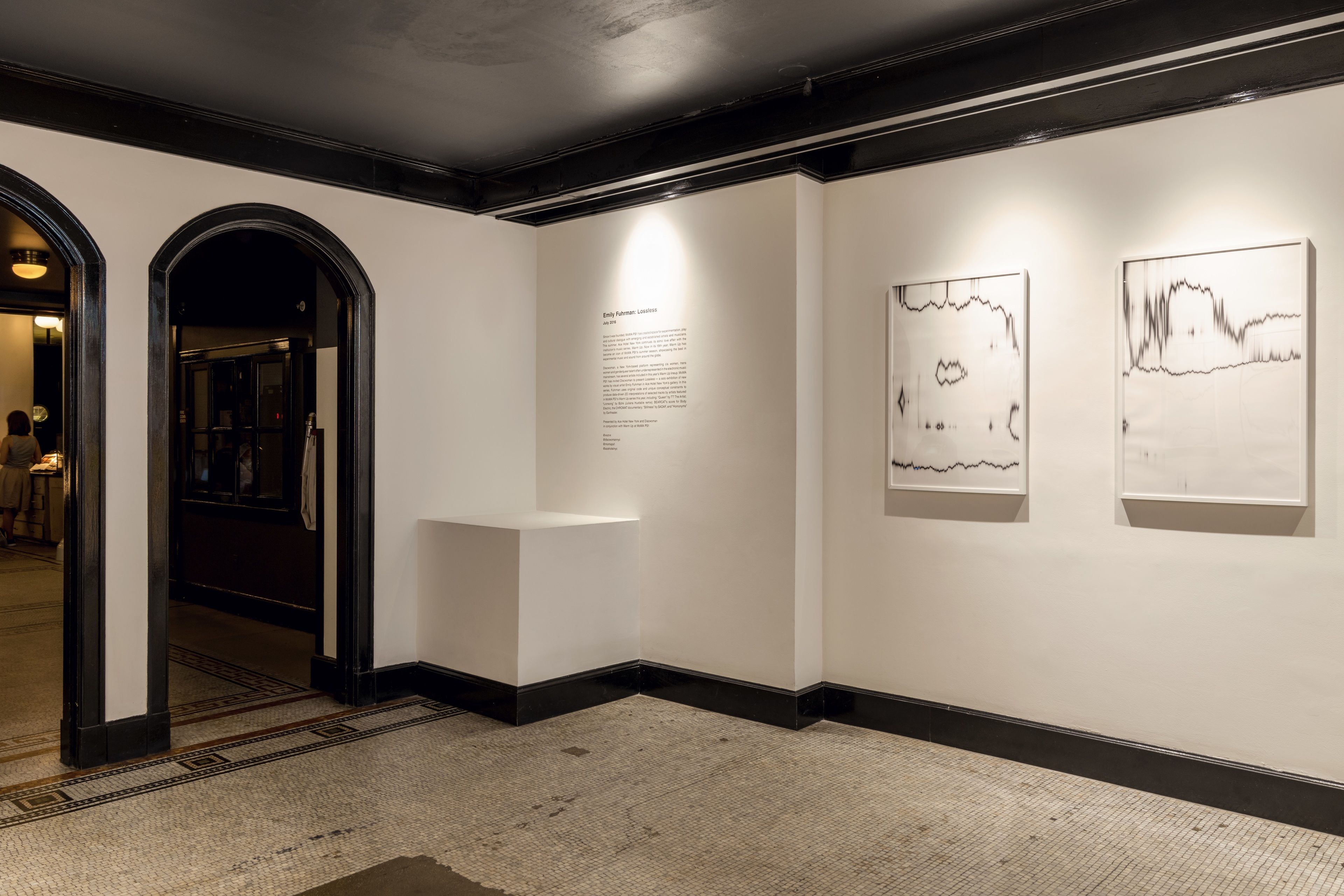 Ein Flur mit weißer Wand, schwarzer Verkleidung und Fliesenboden. An der Wand hängen zwei gerahmte Kunstwerke, und ein kleiner weißer Tisch mit einer Plakette steht daneben.