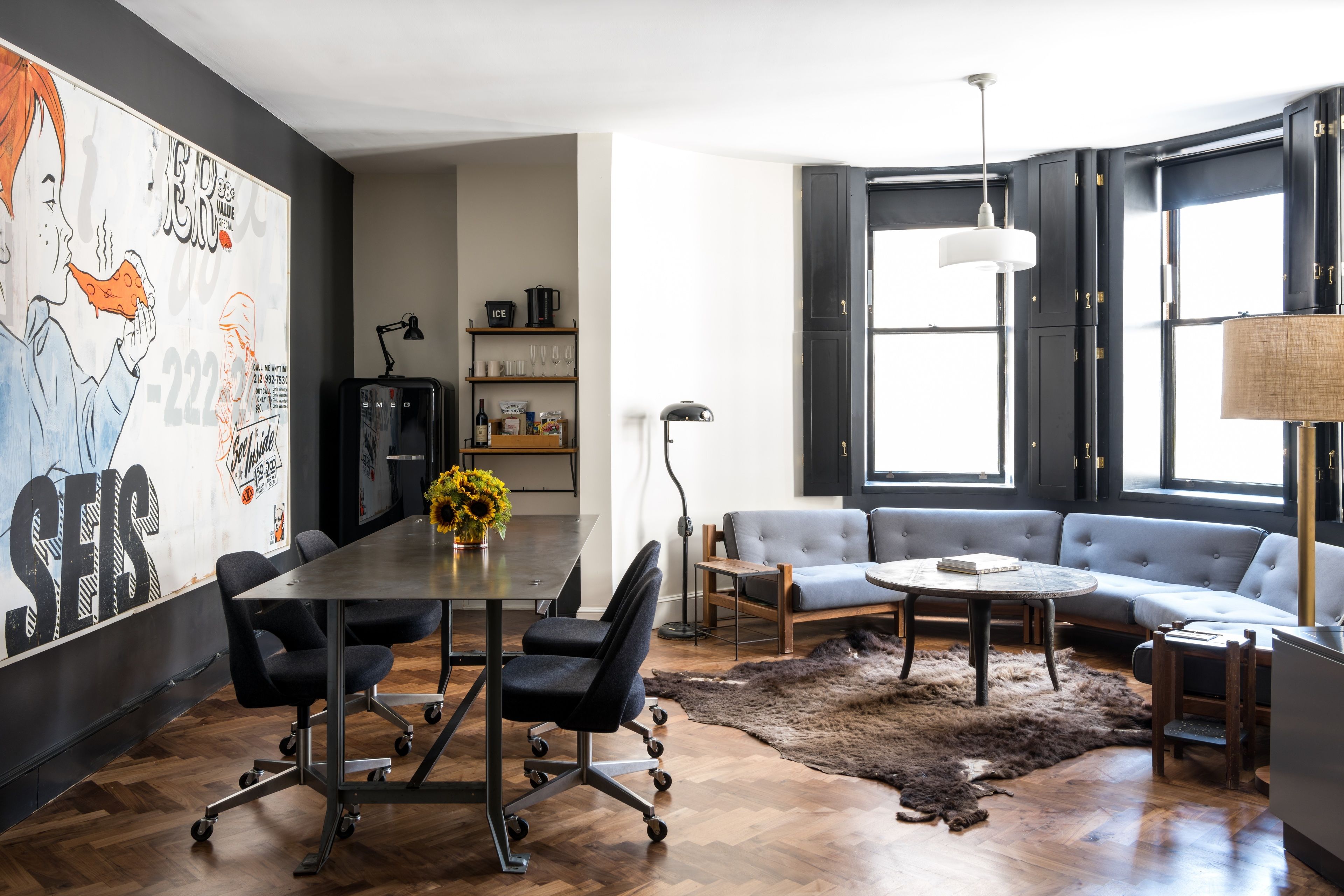 Una sala moderna con piso de madera oscura, un sofá grande, una mesa de centro y una mesa de comedor con sillas. En la pared hay un cuadro abstracto de gran tamaño y una ventana con persianas negras.