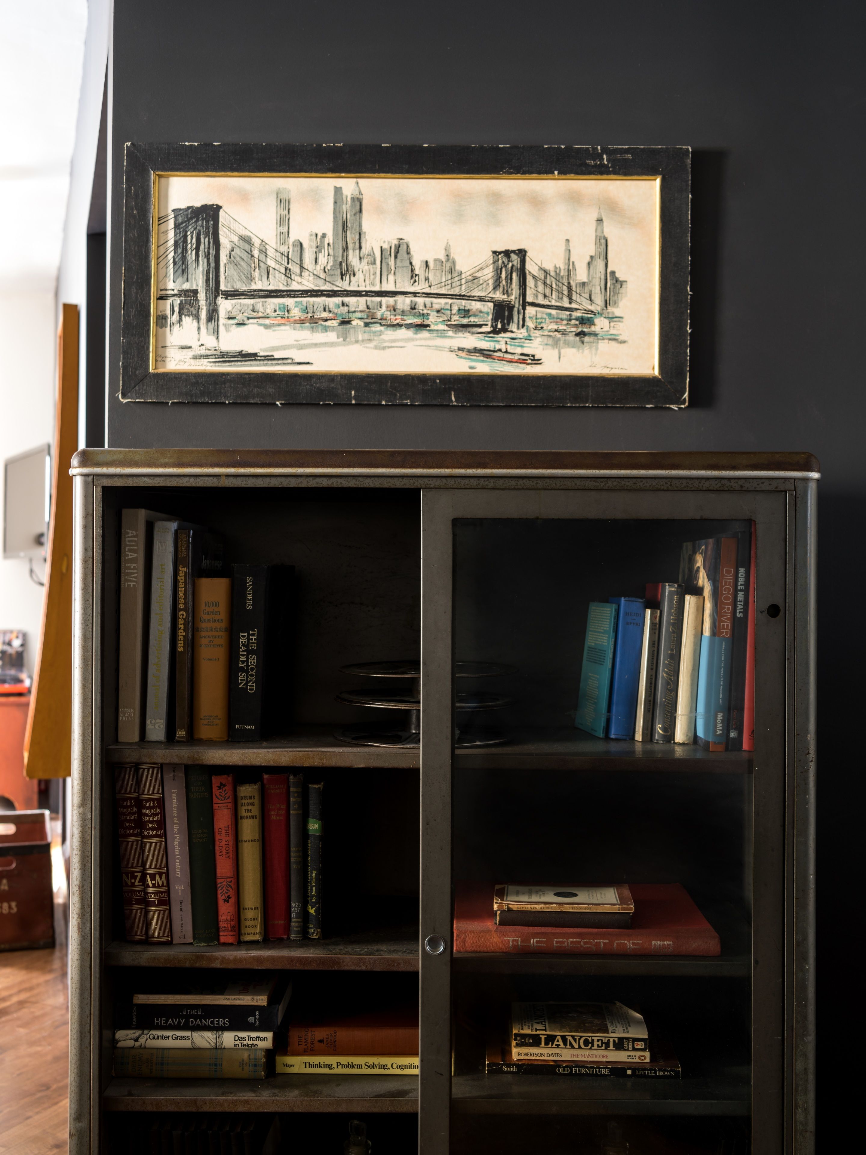 Une bibliothèque remplie de divers livres, et au-dessus, un tableau encadré représentant un paysage urbain.