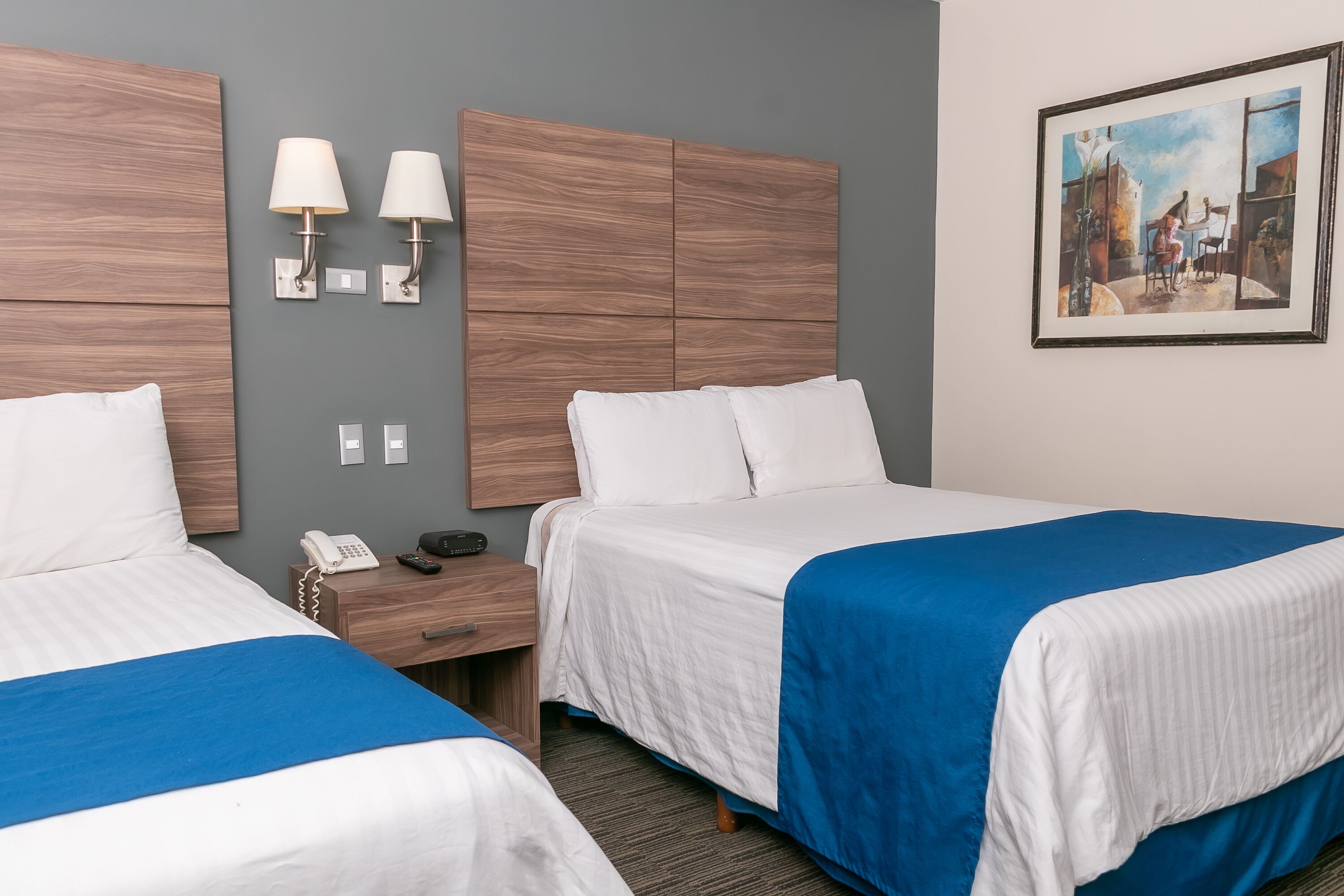 Een hotelkamer met twee bedden, een nachtkastje met telefoon, wandlampen en een schilderij aan de muur.