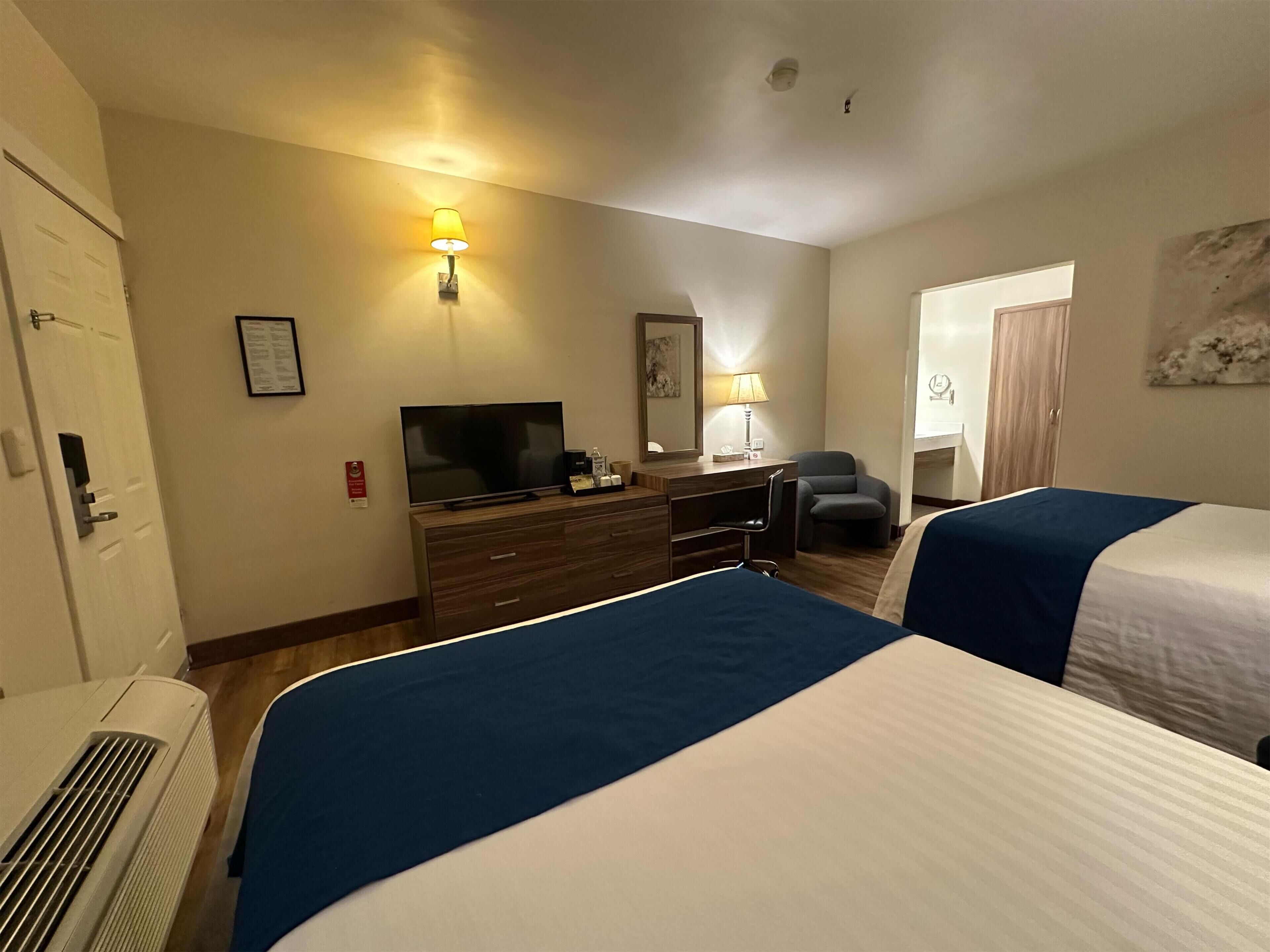 A hotel room with two beds, a desk with a television, a chair, and a lamp.
