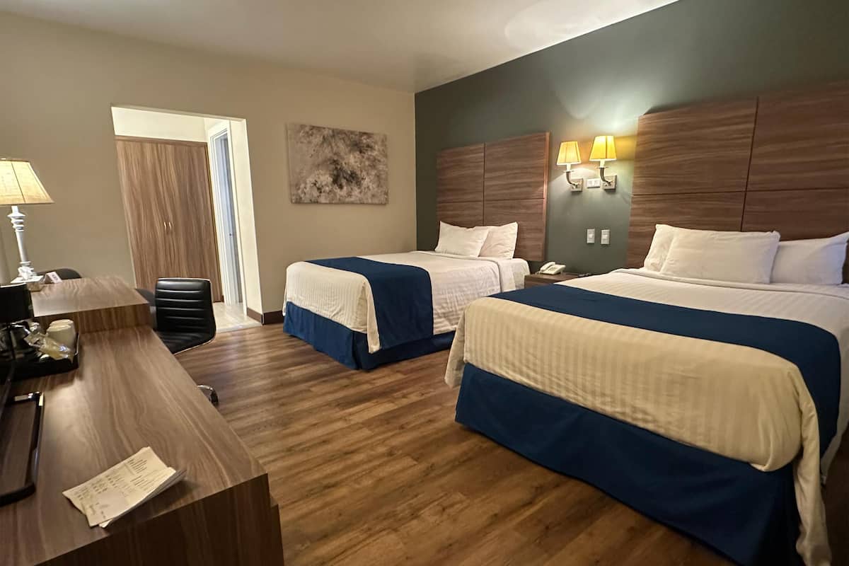 A hotel room with two beds, a desk with a lamp, a telephone, and a painting on the wall.