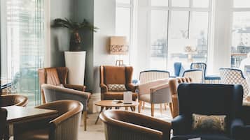 Un área de descanso bien iluminada con sillones de cuero y ratán, una mesa redonda y una gran planta en maceta.