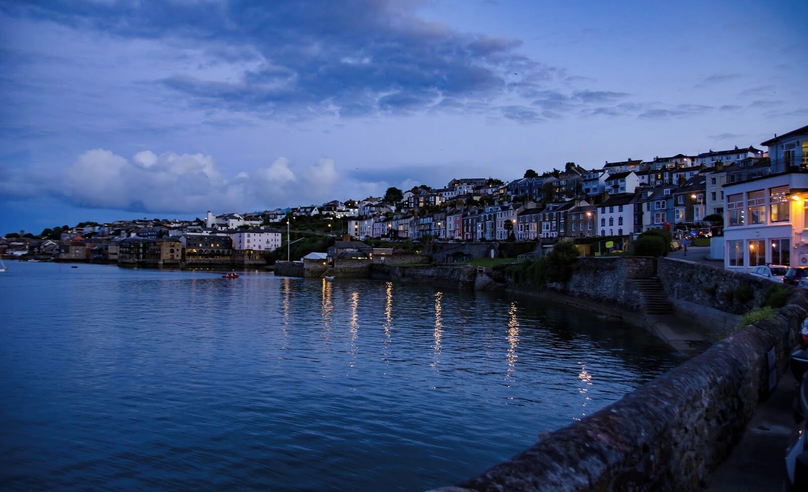 A coastal town at dusk with buildings along the waterfront and a stone wall separating the promenade from the water.