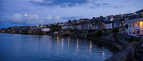 A coastal town at dusk with buildings along the waterfront and a stone wall separating the promenade from the water.