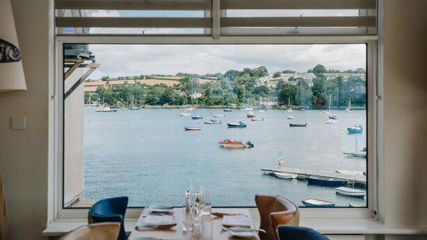 Un'area pranzo con un tavolo apparecchiato per due, con vista su una marina con barche e un paesaggio collinare.