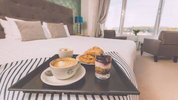 Un lit bien fait, recouvert d’une couverture à rayures, une table d’appoint avec une tasse de café, un croissant et un dessert, dans une chambre d’hôtel.