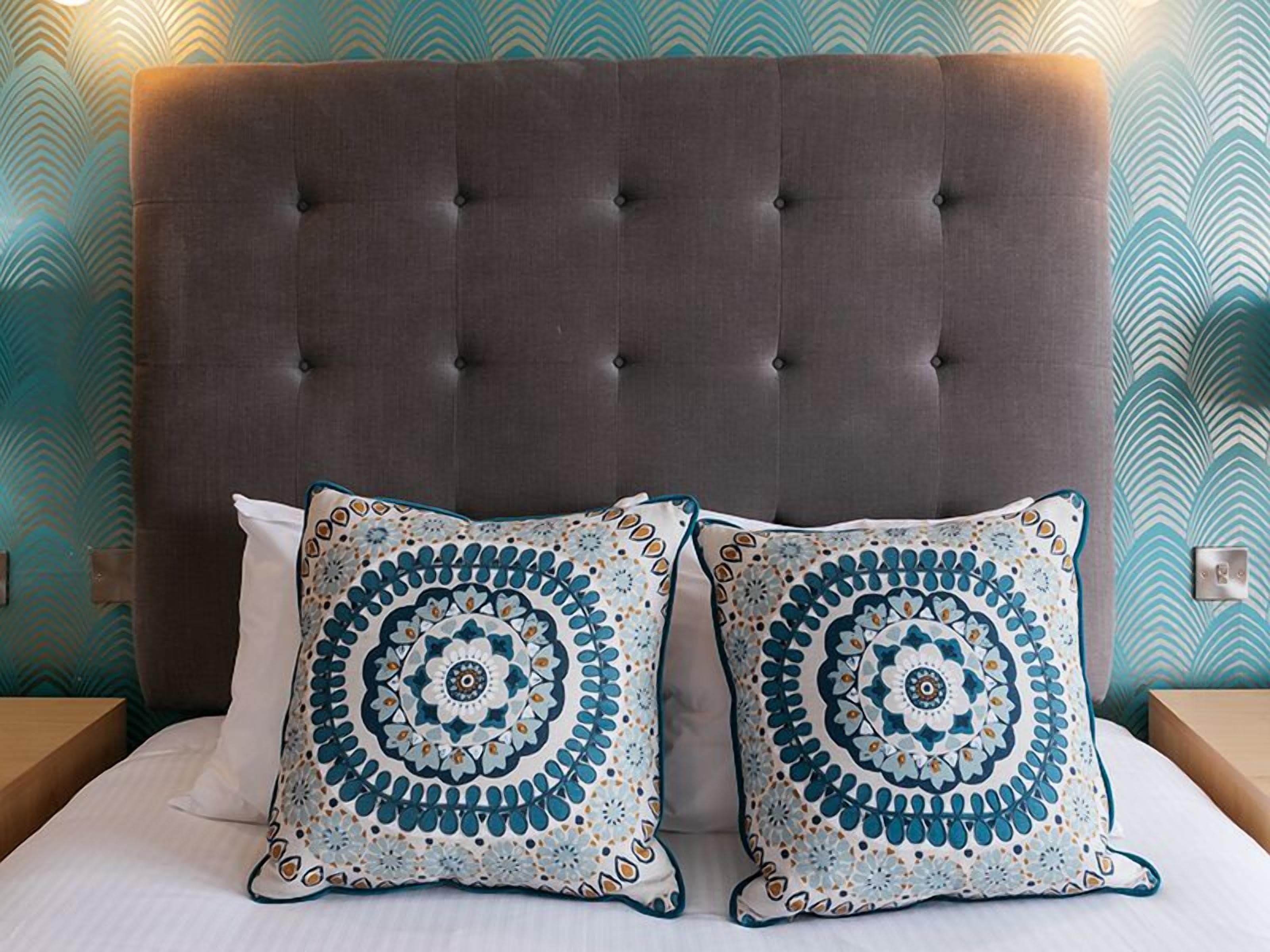 Un lit avec une tête de lit capitonnée, deux coussins à motifs et un mur décoré d’un motif.