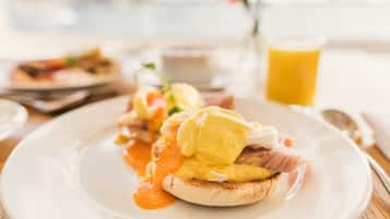 Un plato de desayuno con huevos Benedict, un vaso de jugo de naranja y un florero con una flor roja.