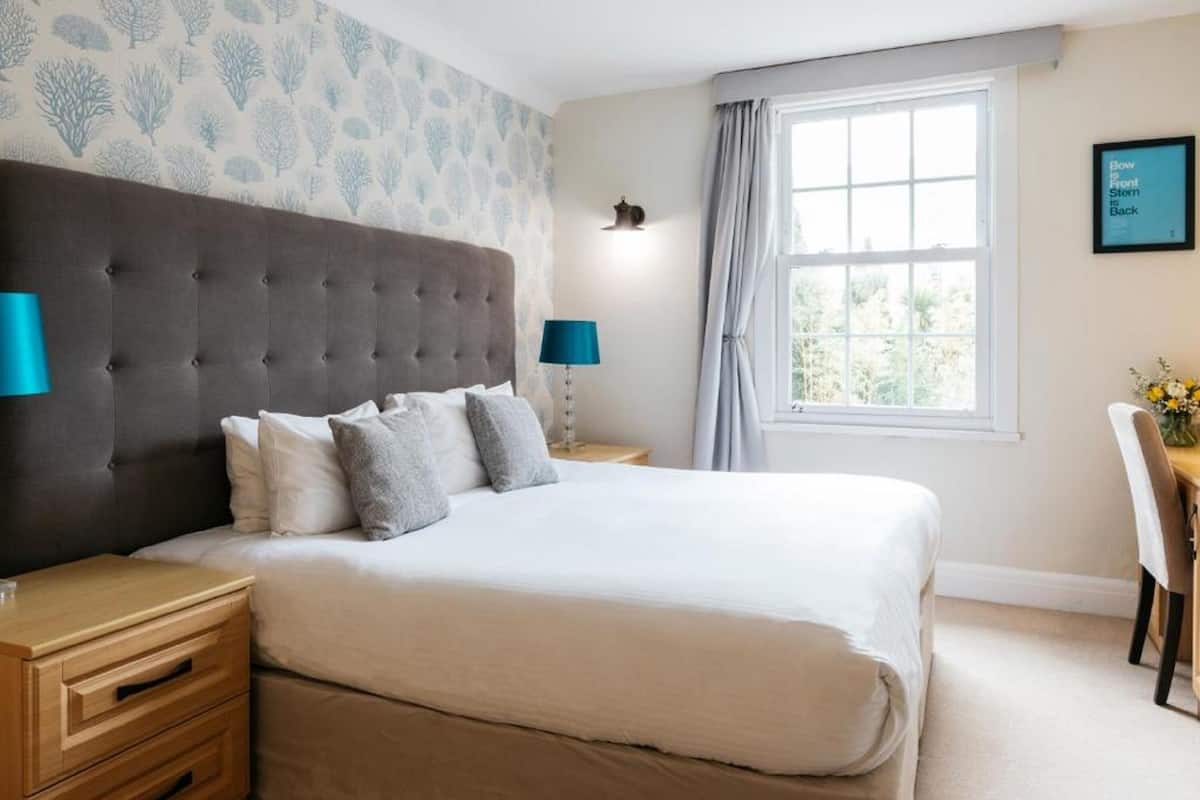 Une chambre à coucher avec une tête de lit capitonnée, un grand lit recouvert de draps blancs, deux tables de chevet avec des lampes, une fenêtre avec des rideaux et un luminaire fixé au mur.