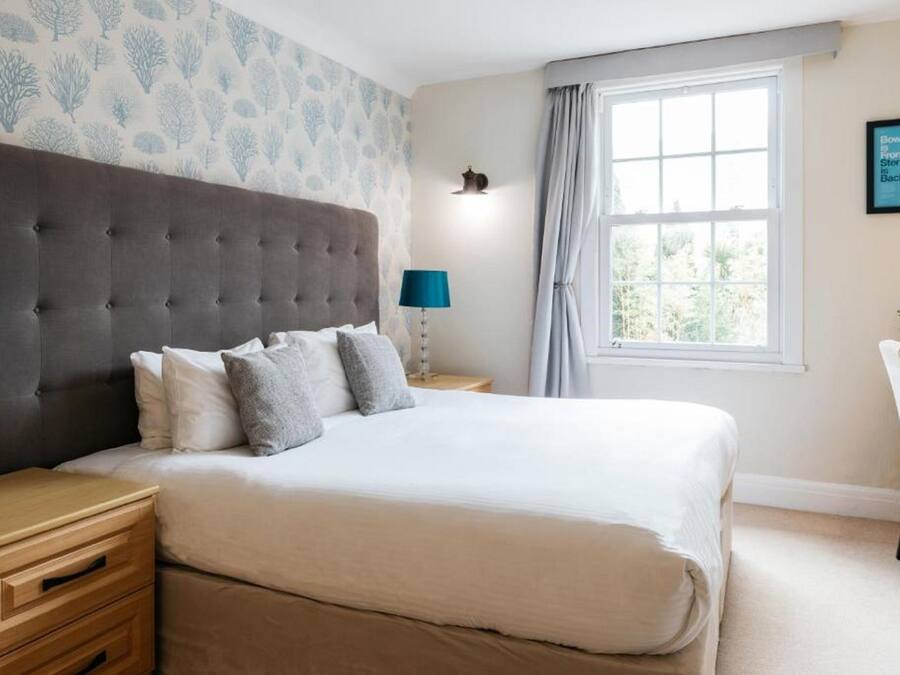 Une chambre à coucher avec une tête de lit capitonnée, un grand lit recouvert de draps blancs, deux tables de chevet avec des lampes, une fenêtre avec des rideaux et un luminaire fixé au mur.