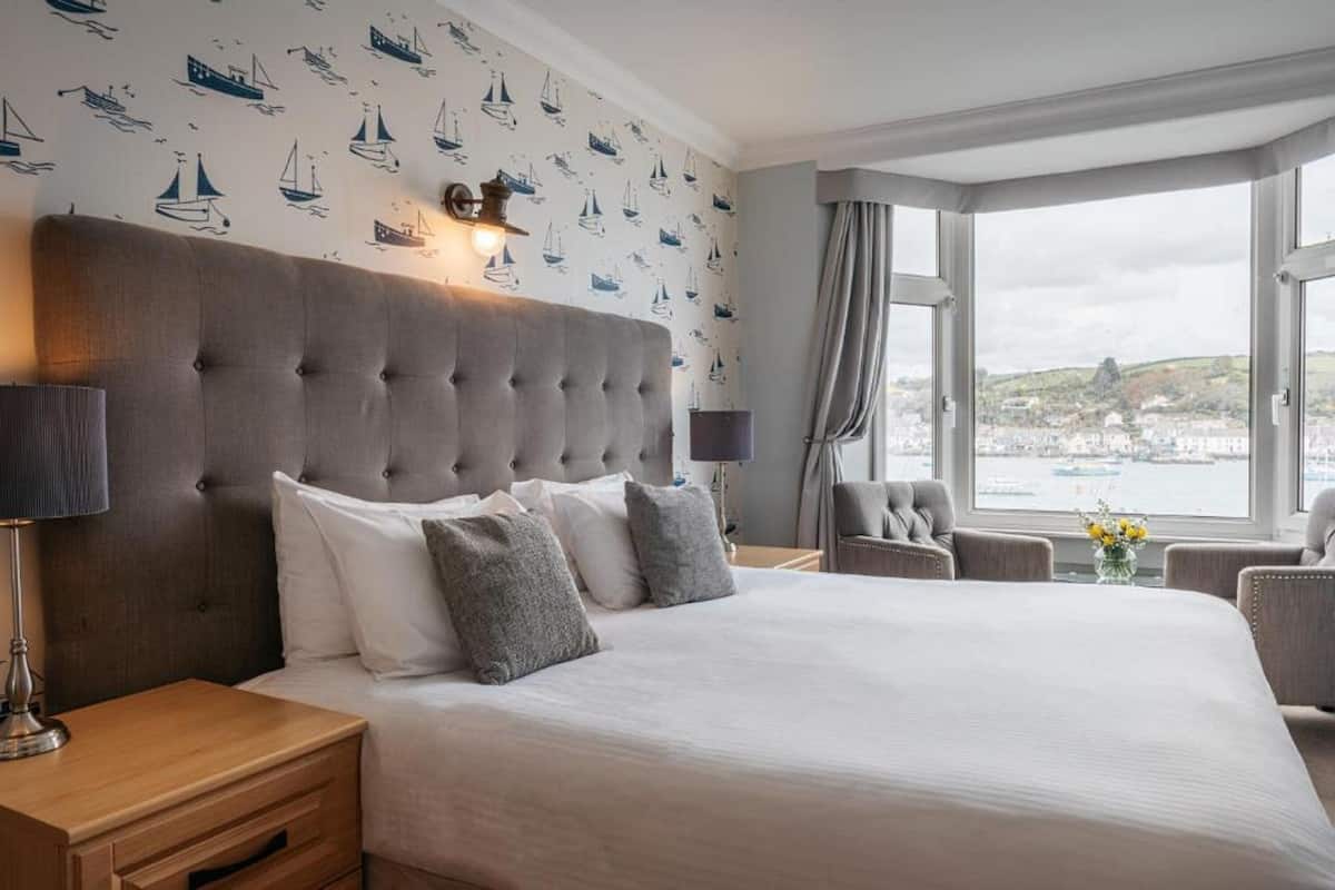 Une chambre d’hôtel dotée d’un grand lit, d’une tête de lit avec un luminaire intégré, d’une vue sur la ville grâce à de grandes fenêtres et d’un mur recouvert d’un papier peint sur le thème de la marine.