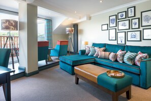 A modern living room with a teal sofa, matching ottoman, and a wooden coffee table. There is a balcony with a view, a floor lamp, and framed artwork on the wall.