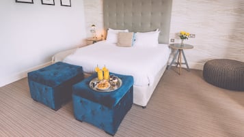 Una camera d'albergo ordinata con un letto grande, due pouf blu, un comodino con una lampada e un tavolino rotondo con un vaso di fiori.