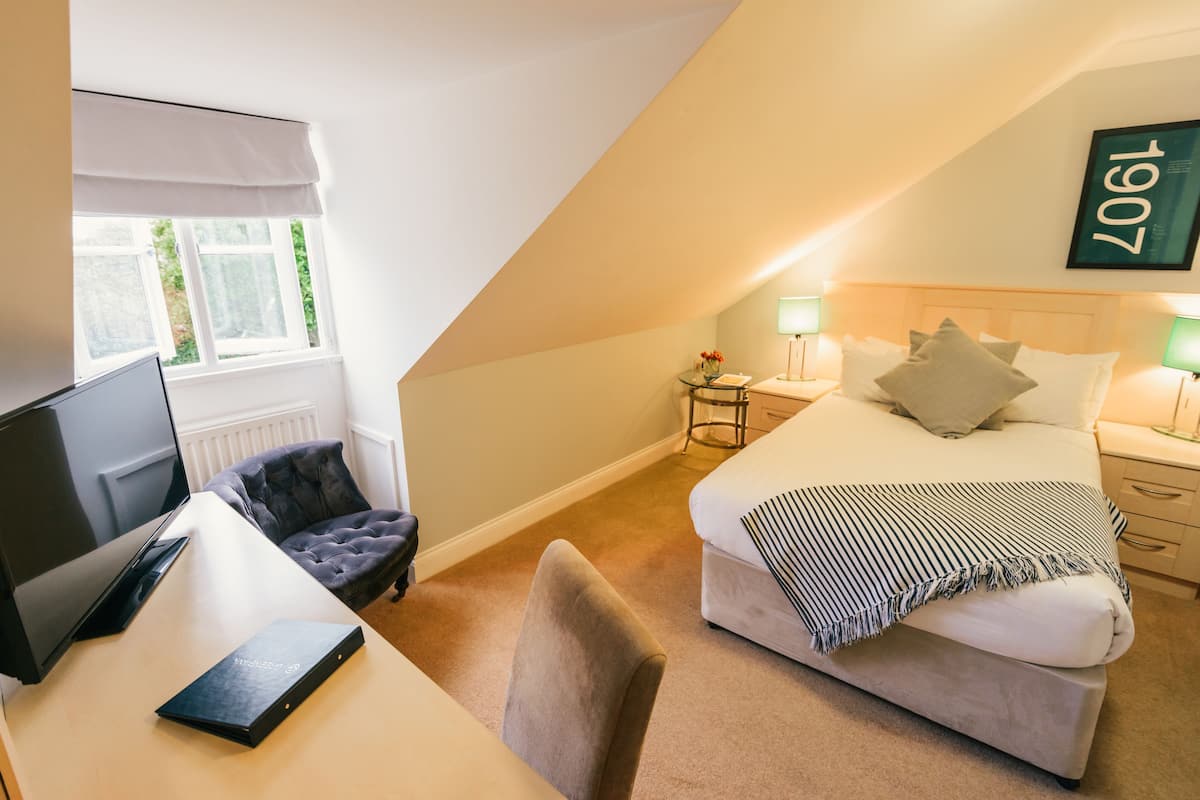 Une chambre à coucher comprenant un lit, des tables de chevet, un bureau avec un ordinateur, une chaise et une fenêtre avec des stores.