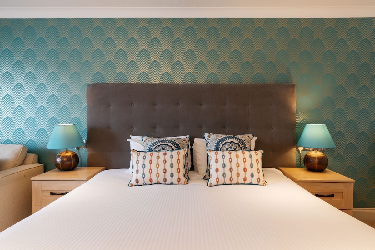 Un lit bien fait, agrémenté de deux coussins décoratifs, une table de chevet en bois avec une lampe, et un mur orné d’un motif.