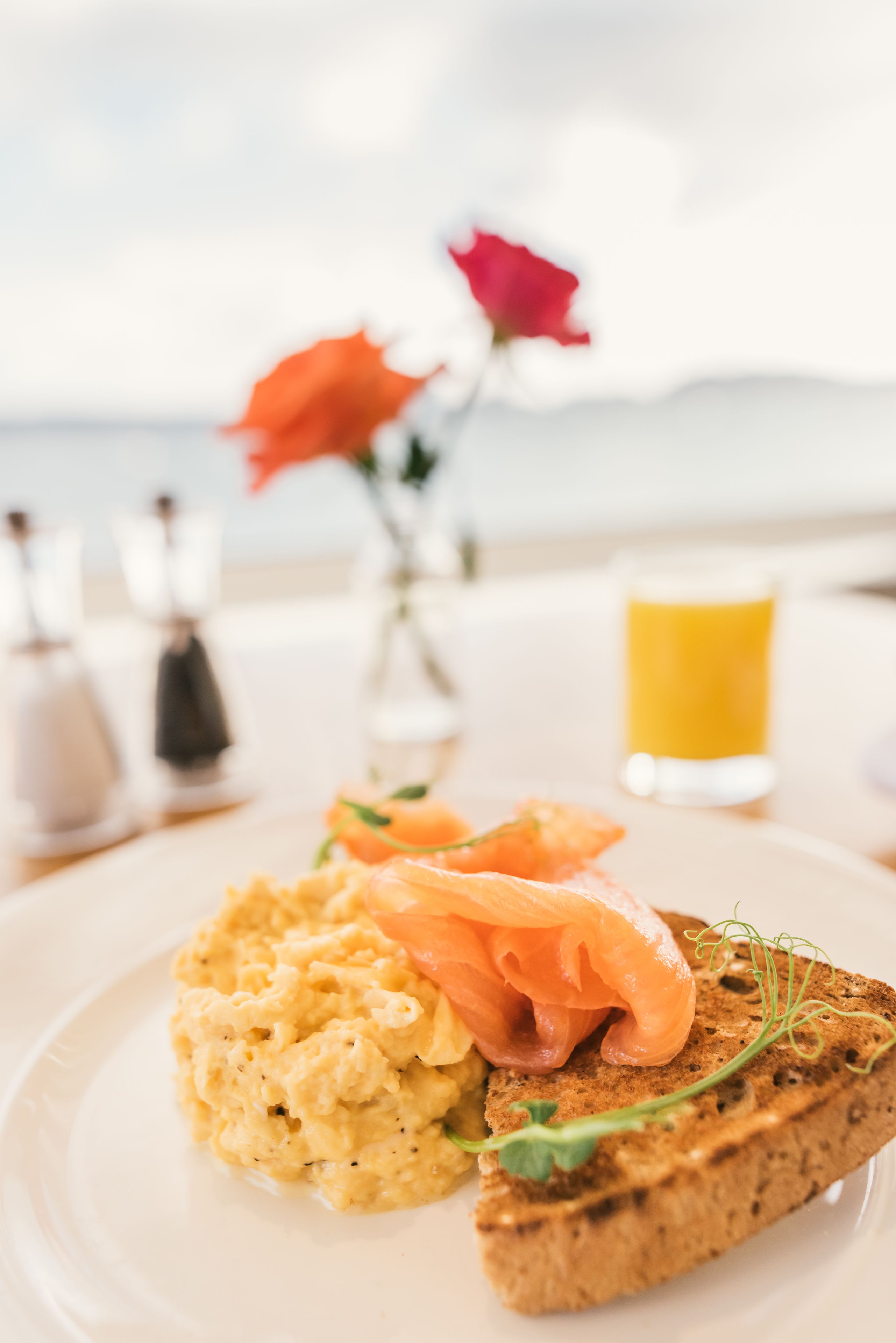 Un piatto con uova strapazzate e salmone affumicato su una fetta di pane, con un bicchiere di succo d'arancia e un vaso di fiori sullo sfondo.