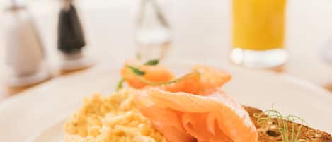 A plate with scrambled eggs and smoked salmon on a slice of bread, with a glass of orange juice and a vase with flowers in the background.