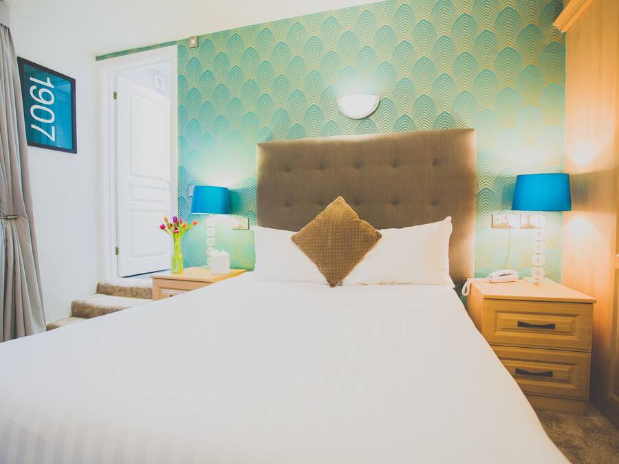 A hotel room with a large bed, two bedside tables with lamps, a nightstand, a wall clock, and a door with a number sign.