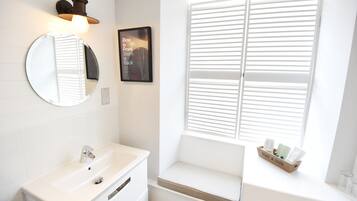 A bathroom with a round mirror, a sink, a window with blinds, and a small bench with toiletries.