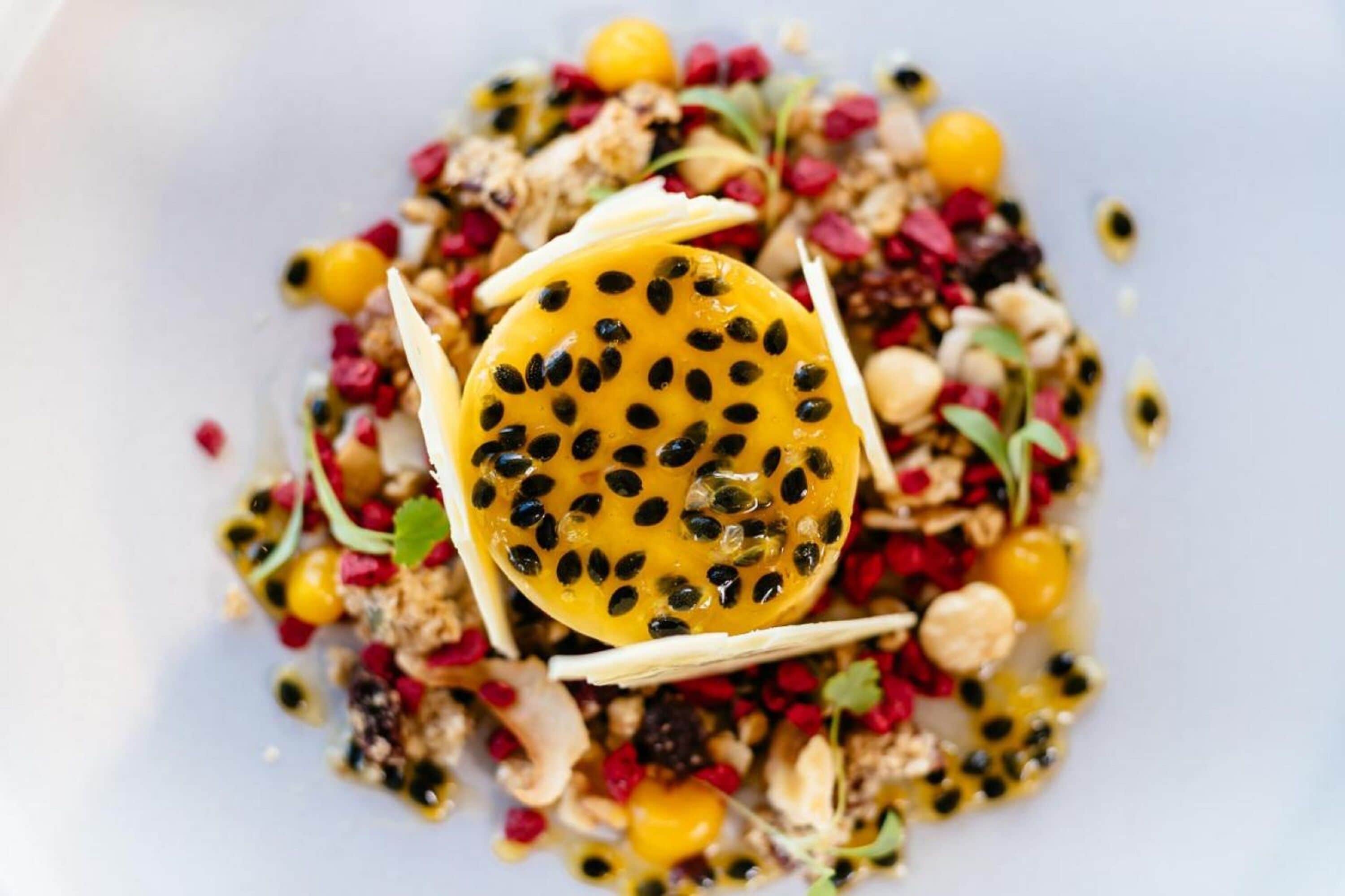 A dish with a slice of yellow fruit topped with black seeds, surrounded by various nuts and seeds.