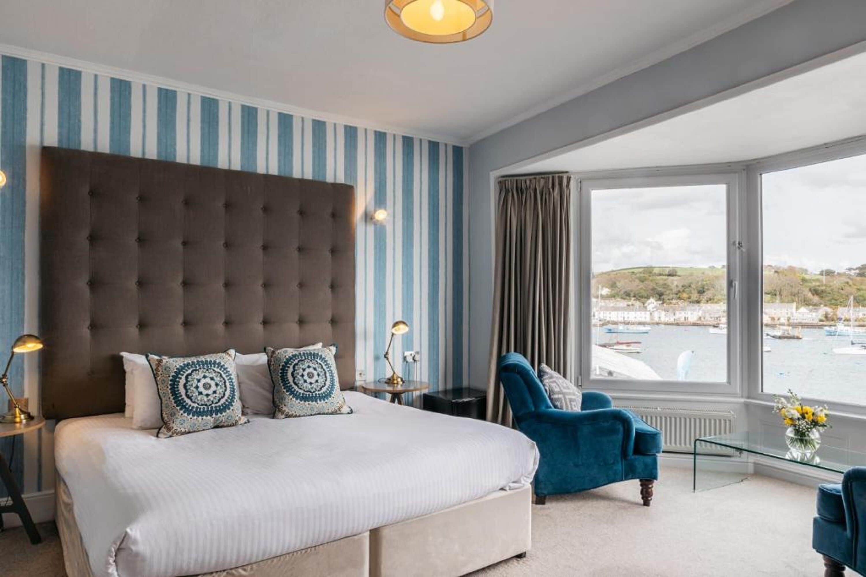 Een slaapkamer met een groot bed, een gestoffeerd hoofdeinde, uitzicht op zee en een blauwe fauteuil.