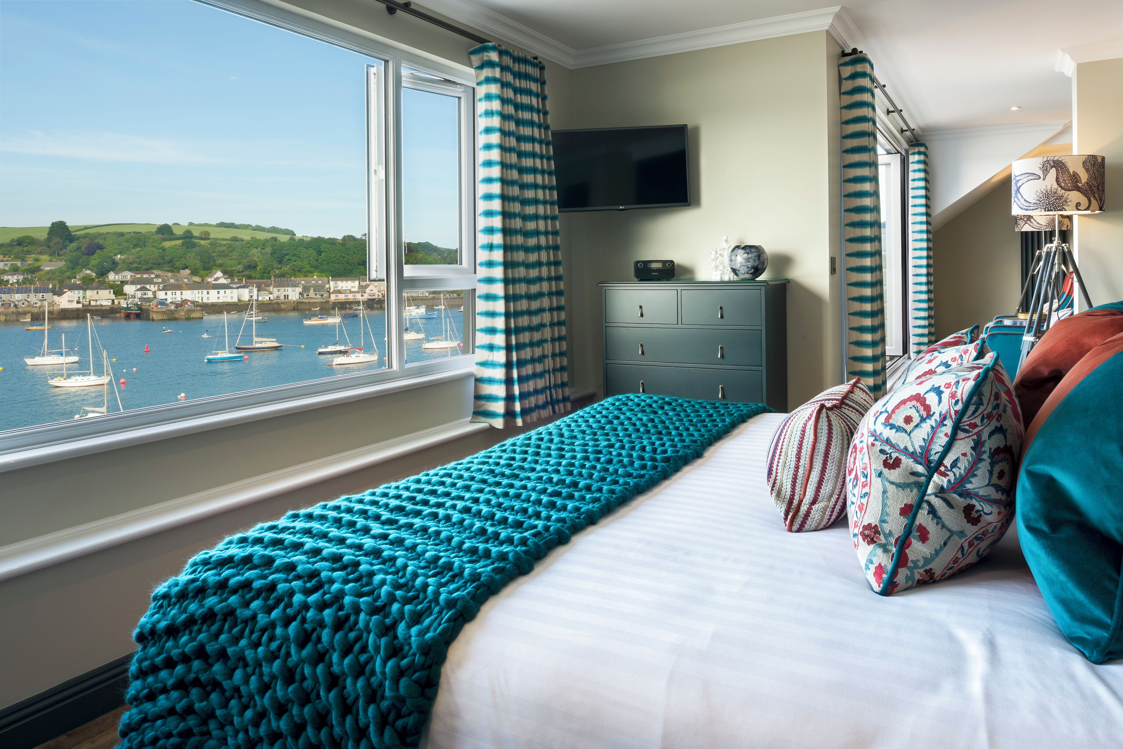 A bedroom with a large window overlooking a harbor, a bed with teal bedding, and a dresser with a flat-screen TV.