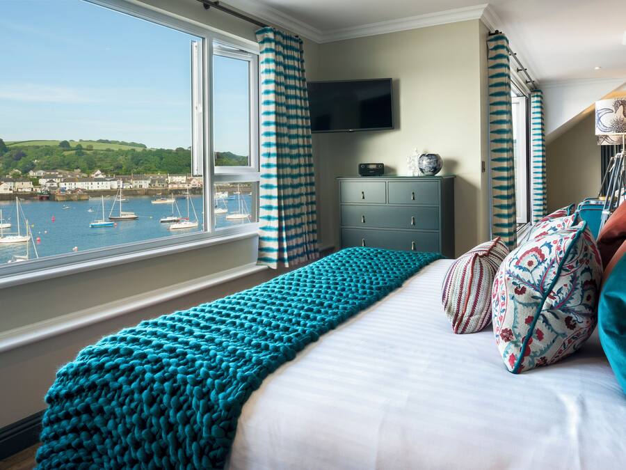 A bedroom with a large window overlooking a harbor, a bed with teal bedding, and a dresser with a flat-screen TV.