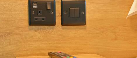 A remote control on a wooden table with a wall-mounted electrical outlet and switch.
