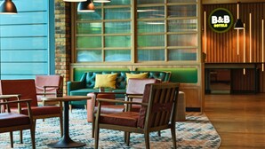 Un hall de hotel con un sofá verde, sillas de madera, alfombra estampada, iluminación colgante y un amplio ventanal con persianas.