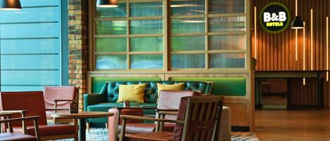 A hotel lobby with a green sofa, wooden chairs, a patterned rug, pendant lighting, and a large window with blinds.