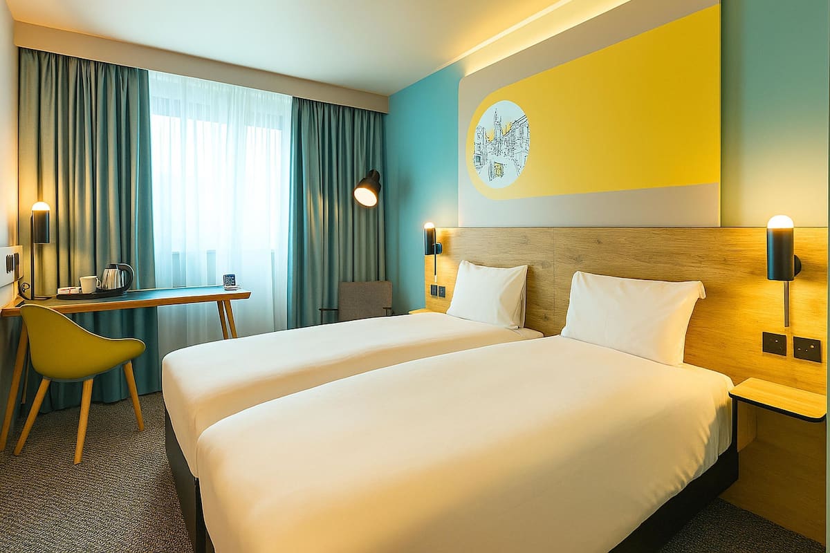 A hotel room with two beds, a desk, a chair, and a large window with curtains.