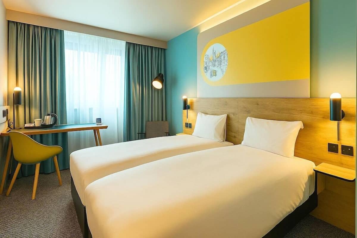 A hotel room with a large bed, a desk, a chair, and a wall-mounted lamp.