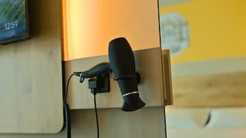 Un secador de pelo negro conectado a un enchufe de pared, con un panel de madera y una pared amarilla al fondo.