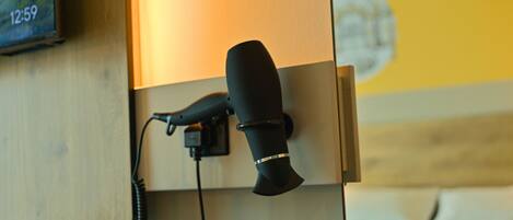 A black hair dryer plugged into a wall socket, with a wooden panel and a yellow wall in the background.