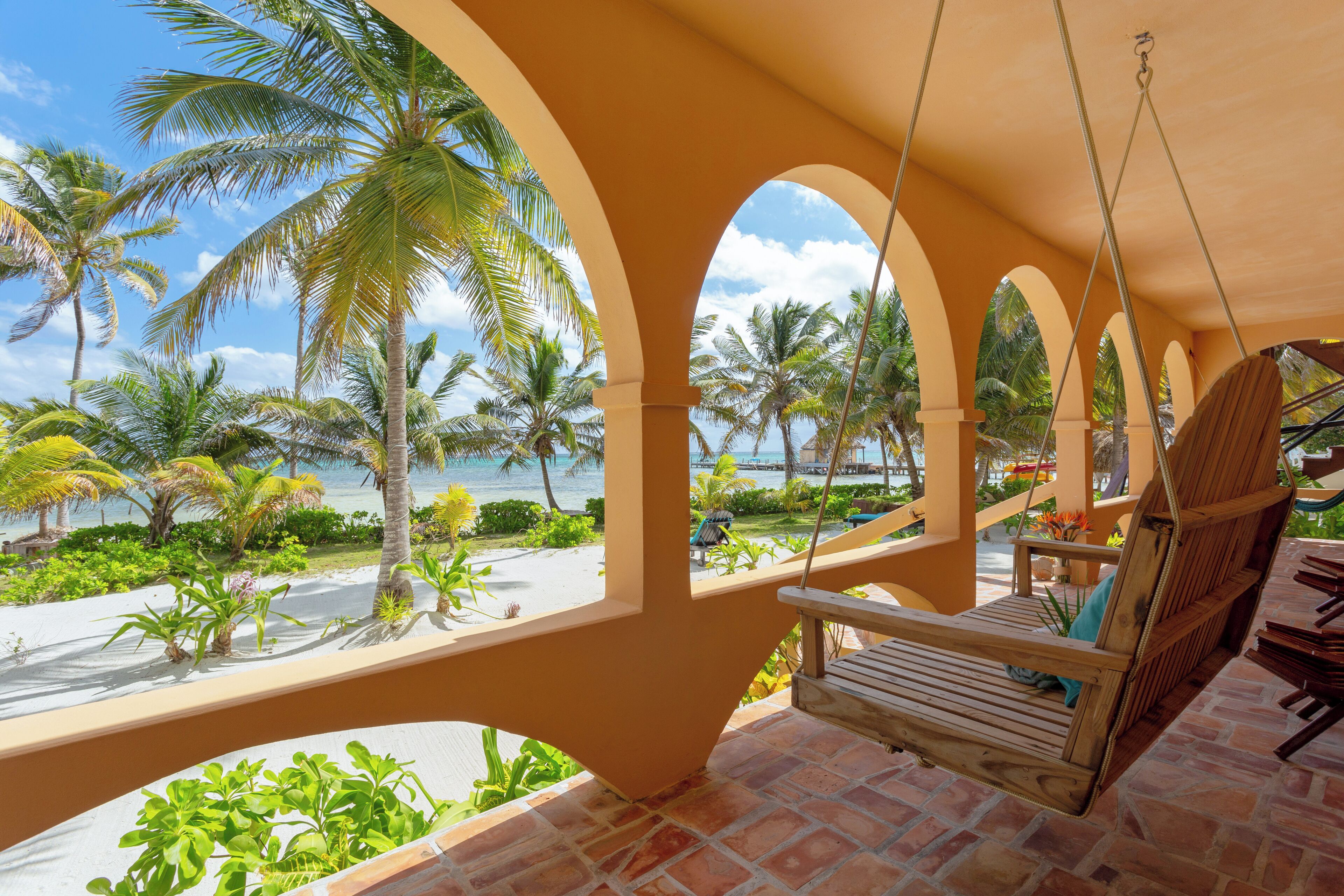 A wooden swing chair hanging from a structure with arches, overlooking a beach with palm trees and the ocean.