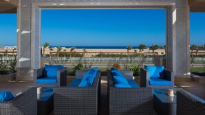 Un espace extérieur aménagé avec des meubles en osier et des coussins bleus, protégé par un auvent, et offrant une vue sur la plage et la piscine.