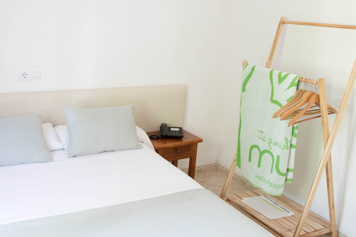 Een opgemaakt bed met kussens, een nachtkastje met een telefoon, en een houten kledingrek met kleding.