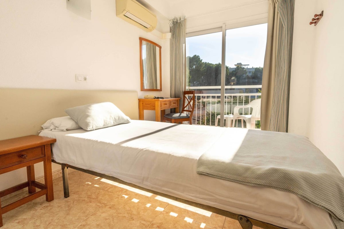 Een slaapkamer met een bed, een houten nachtkastje, een bureau, een stoel en een balkon met uitzicht.