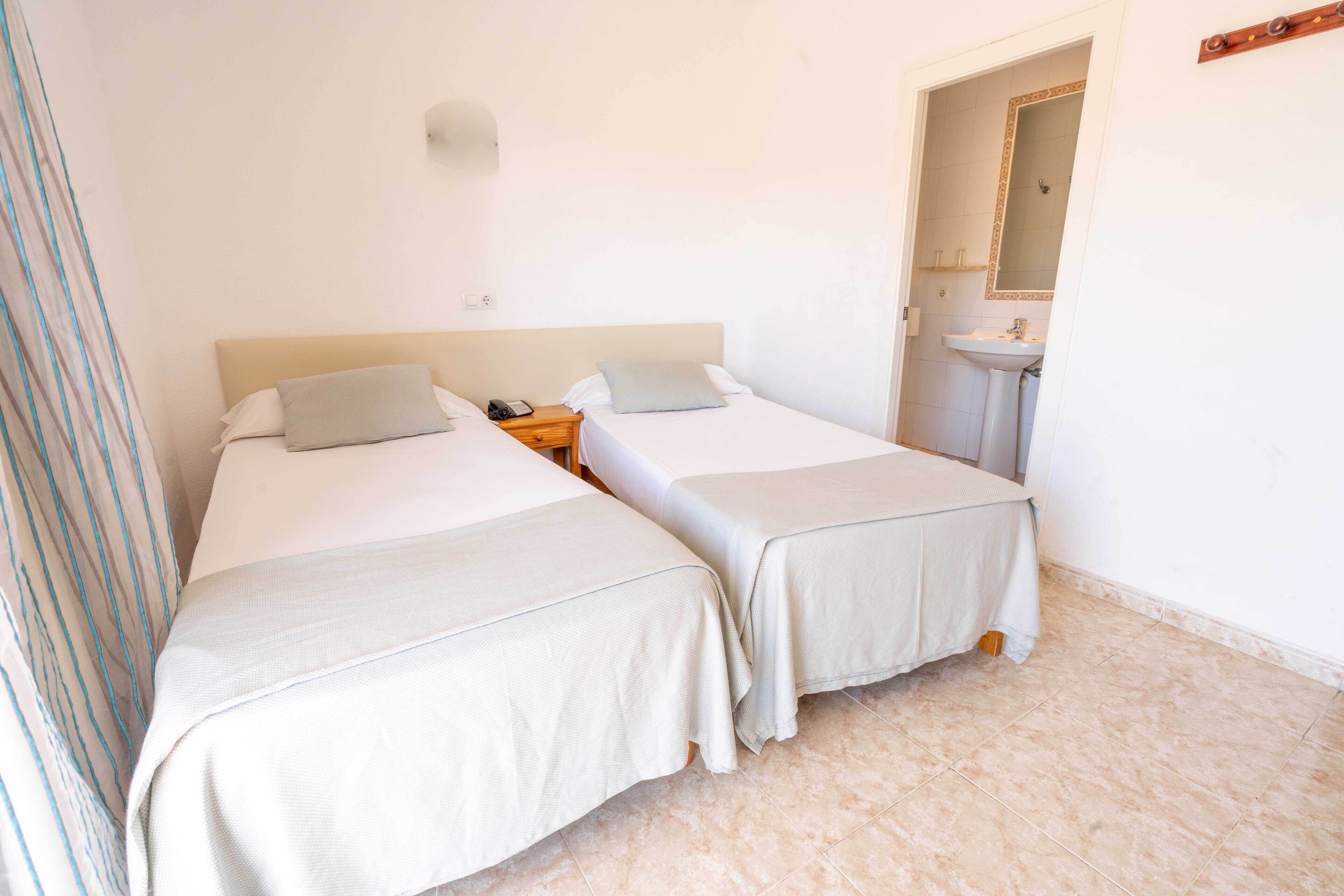 A room with two beds, a bedside table with a lamp, a mirror, and a bathroom with a sink and mirror.