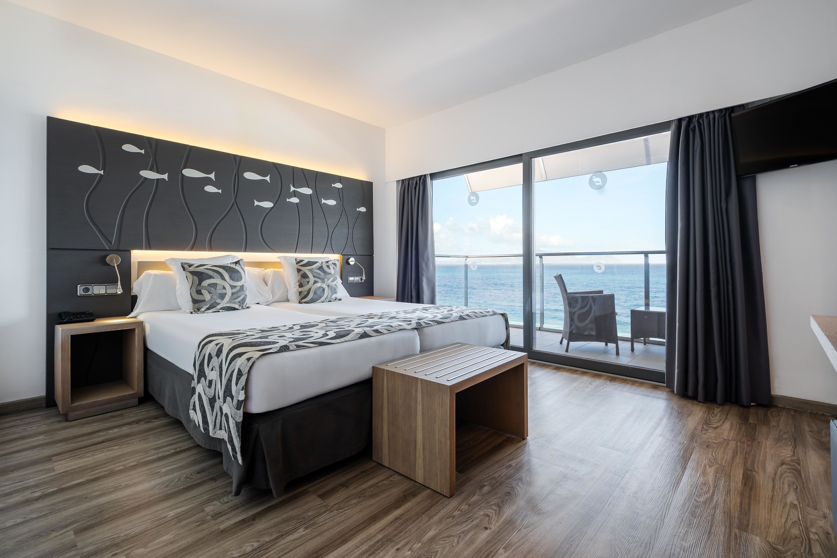 Ein modernes Hotelzimmer mit einem großen Bett, Nachttischen, einer Bank und Meerblick durch ein großes Fenster.