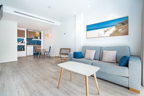A modern living room with a grey sofa, a white coffee table, and a dining area with a wooden table and chairs. - THB Gran Bahia Hotel and Apartments (Santa Margalida)