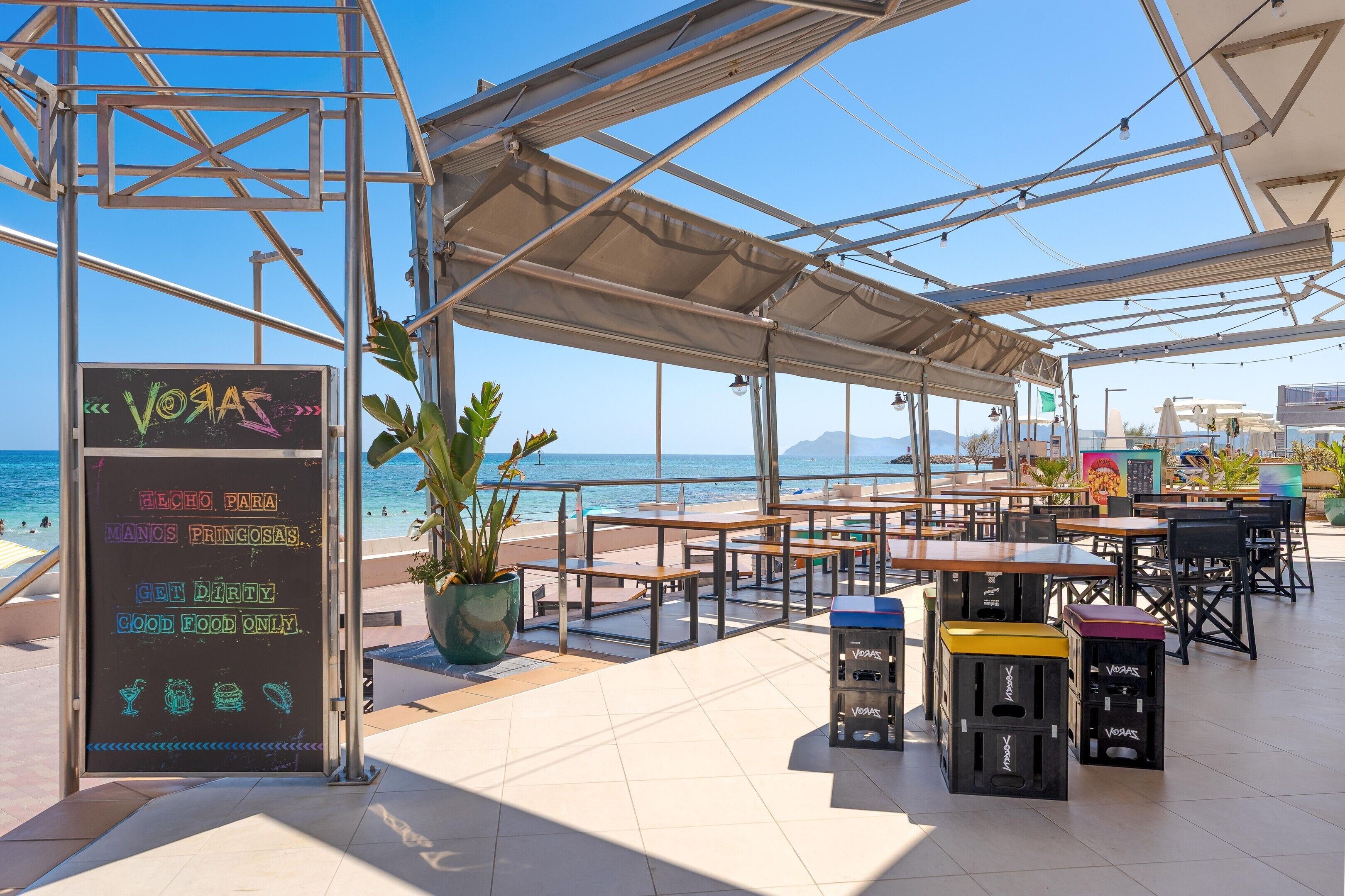 Un espacio al aire libre con mesas y sillas, con vista a una playa y al mar.