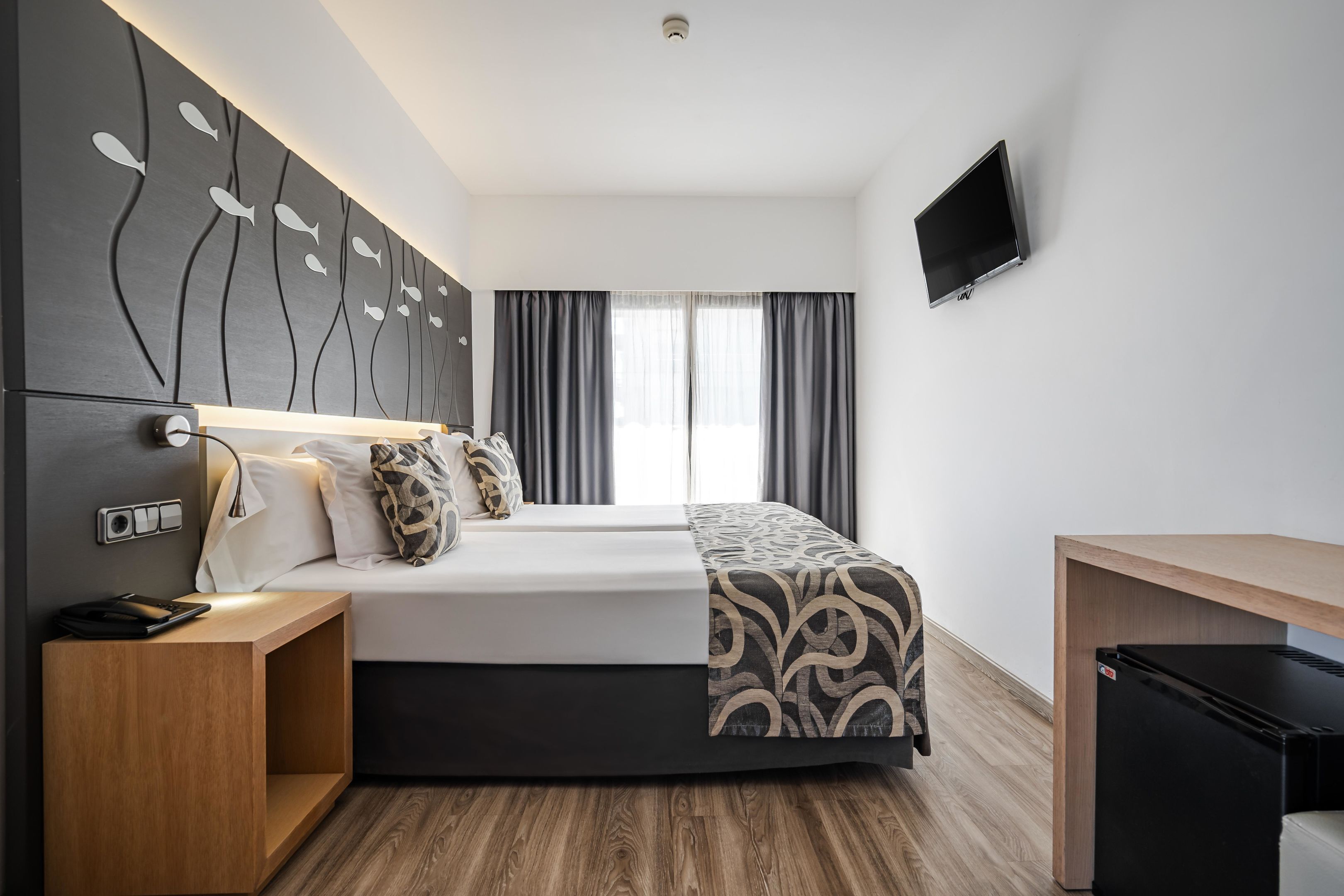 A modern hotel room with a bed, bedside table, TV, and a small refrigerator.