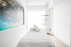 A minimalist hotel room with a bed, a glass cabinet, and a large painting of a tropical scene.