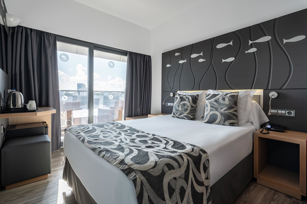 Une chambre d’hôtel moderne dotée d’un grand lit, d’un bureau avec une bouilloire et offrant une vue sur la ville grâce à de grandes fenêtres.