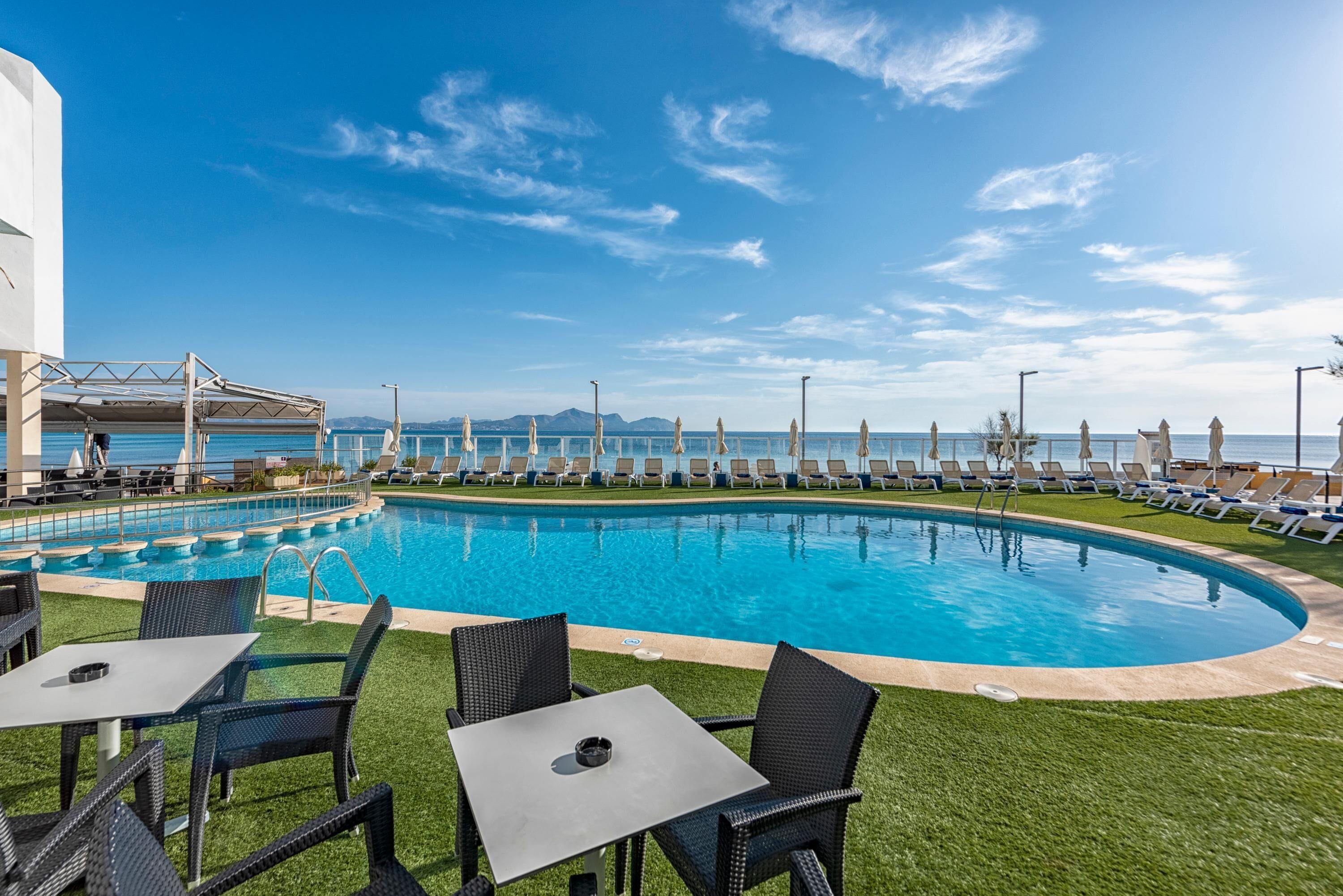 A pool area with lounge chairs and tables, a clear sky, and a view of the sea.