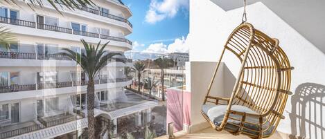 A balcony with a wicker chair and a view of a multi-story building with balconies and palm trees.