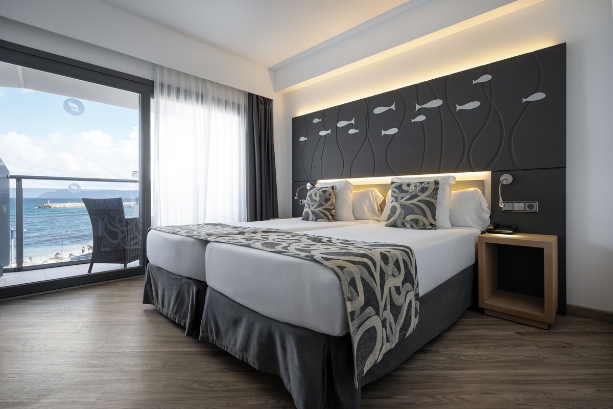 Une chambre d’hôtel moderne avec un grand lit, une table de chevet et une vue sur la mer par une grande fenêtre.